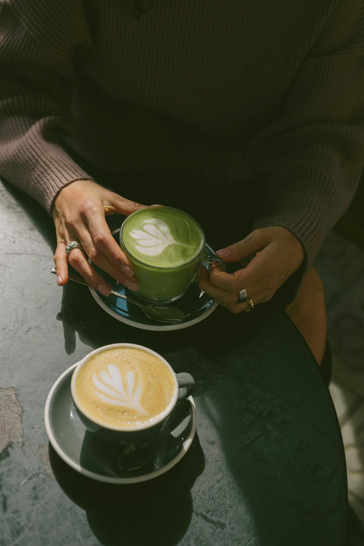 Mains tenant un matcha latte vert avec un golden latte doré posé sur la table