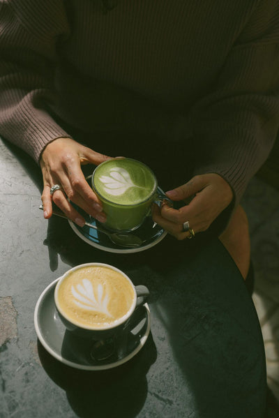 Mains tenant un matcha latte vert avec un golden latte doré posé sur la table